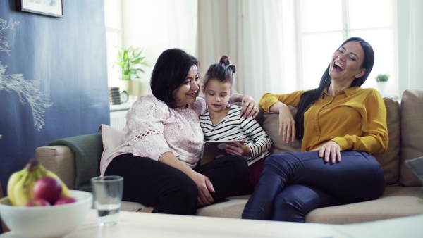 A happy small girl with mother and grandmother sitting on sofa at home, using tablet.