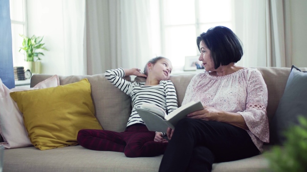 A small girl with grandmother sitting on sofa at home, reading a story book.