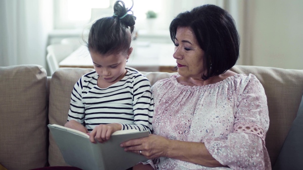 A small girl with grandmother sitting on sofa at home, reading book together.