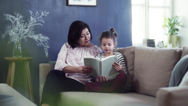 A small girl with grandmother sitting on sofa at home, reading book together.