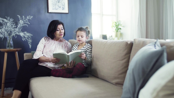 A small girl with grandmother sitting on sofa at home, reading book together.