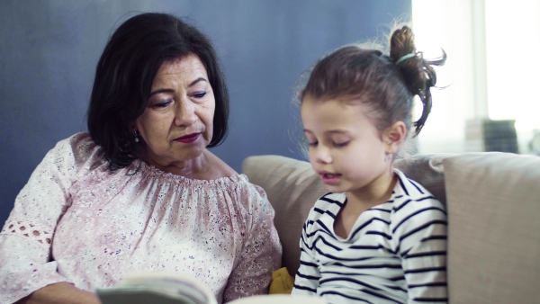 A small girl with grandmother sitting on sofa at home, reading book together.