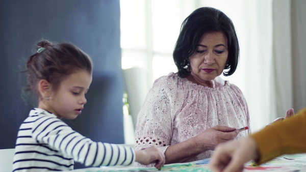 A small girl with grandmother sitting at the table at home, painting pictures.