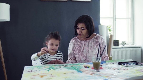 A small girl with grandmother sitting at the table at home, painting pictures.