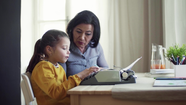 A happy small girl with grandmother sitting at table at home, using typewriter.