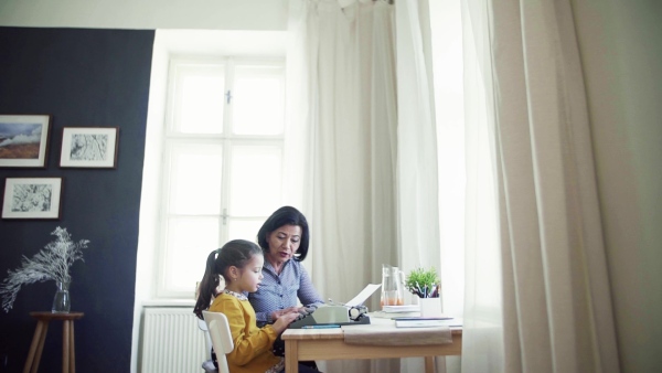 A happy small girl with grandmother sitting at table at home, using typewriter. Slow motion.