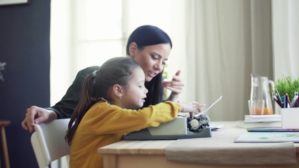 A happy small girl with mother sitting at table at home, using typewriter.