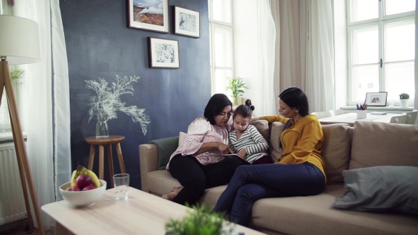 A small girl with mother and grandmother sitting on sofa at home, using tablet. Slow motion.