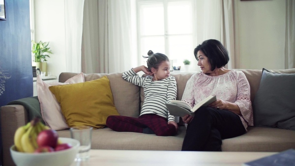 A small girl with grandmother sitting on sofa at home, reading book together. Slow motion.