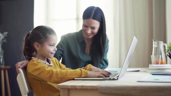 A happy small girl with mother sitting at table at home, using laptop.