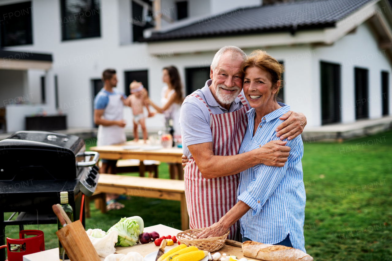 Front view of senior couple with family outdoors on garden barbecue, grilling.