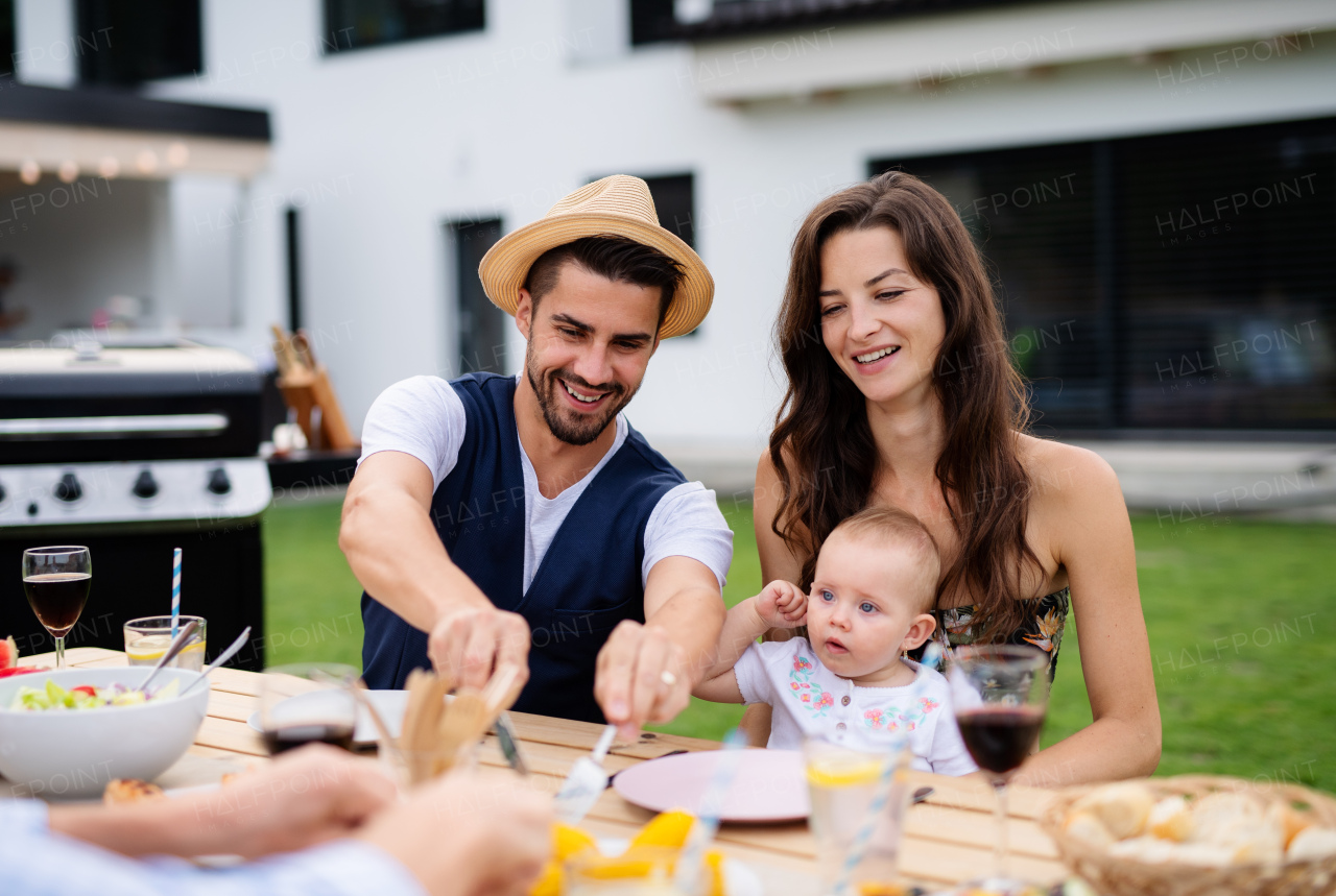 Young couple with baby sitting at table outdoors on family garden barbecue, eating.