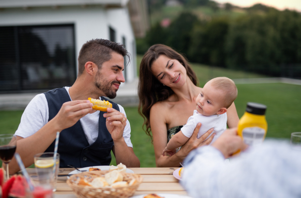 Young couple with baby sitting at table outdoors on family garden barbecue, eating.