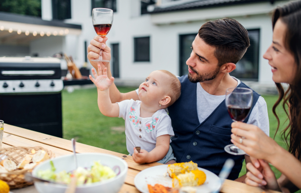 Young couple with baby sitting at table outdoors on family garden barbecue, drinking wine.