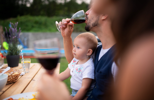 A young couple with baby sitting at table outdoors on family party, drinking wine.