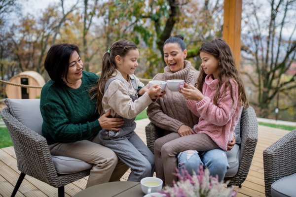 Two happy sisters with a mother and grandmother sitting and drinking tea outdoors in patio in autumn.