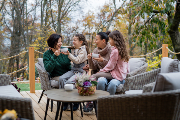 Two happy sisters with a mother and grandmother sitting and drinking tea outdoors in patio in autumn.