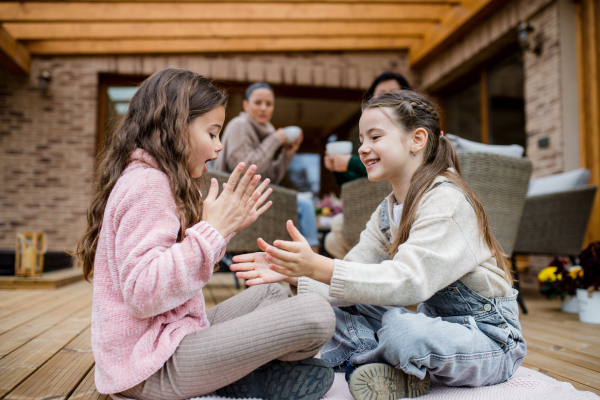 Two happy little sisters hugging outdoors in a patio in autumn, mother and grandmother at background.