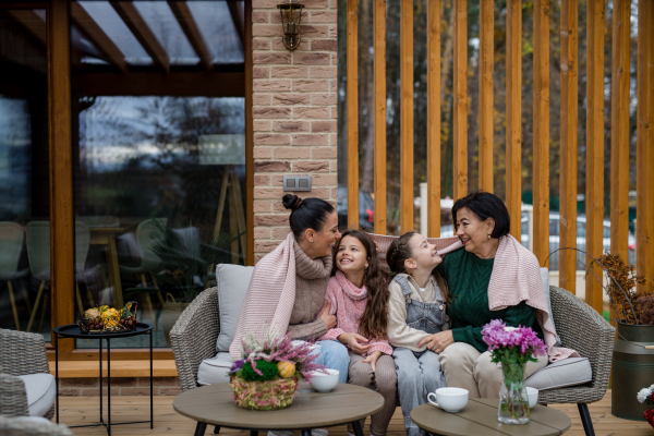 Two happy sisters with a mother and grandmother sitting wrapped in blanket outdoors in patio in autumn