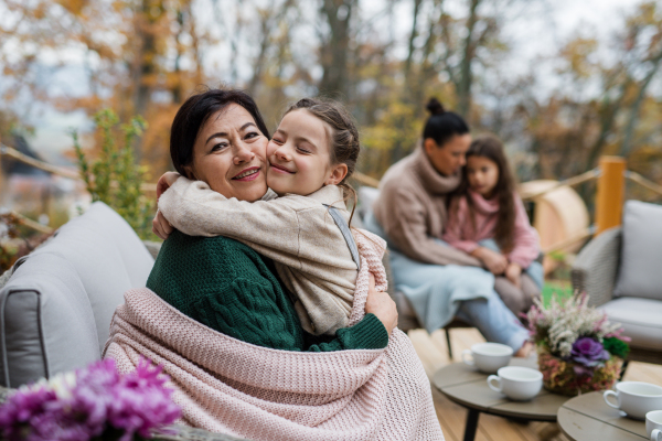 A happy little girl with grandmother sitting together wrapped in blanket outdoors in patio in autumn