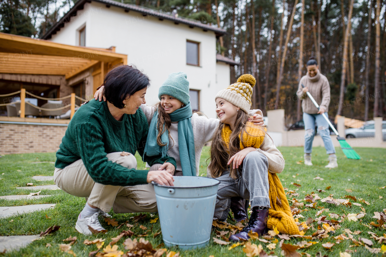 Happy little girls with a grandmother picking up leaves and putting them in bucket in garden in autumn