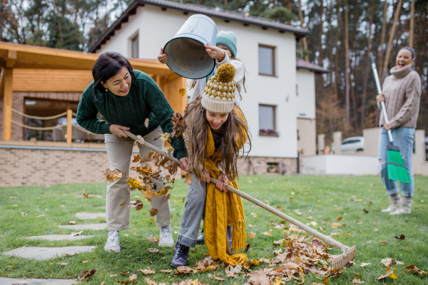 Happy little girls with a grandmother picking up leaves and putting them in bucket in garden in autumn