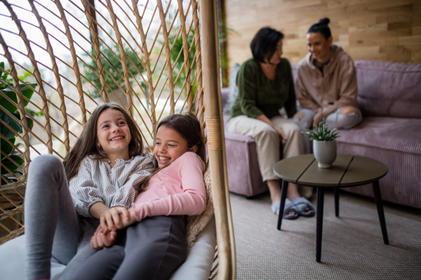 Happy little sisters sitting in wicker rattan hang chair indoors in conservatory, with mother and grandmother at background.