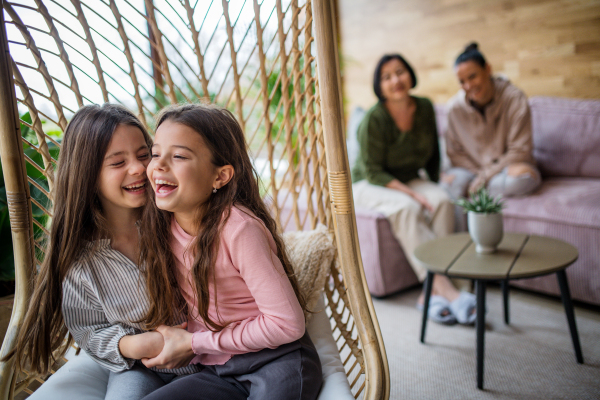 Happy little sisters sitting in wicker rattan hang chair indoors in conservatory, with mother and grandmother at background.