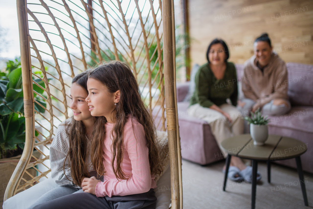 Happy little sisters sitting in wicker rattan hang chair indoors in conservatory, with mother and grandmother at background.