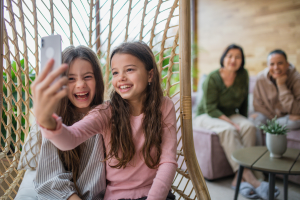 Happy little sisters sitting in a wicker rattan hang chair indoors in conservatory and taking selfie.