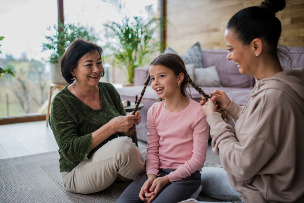 A happy little girl sitting on floor, her mother and grandmother making her plaits at home.