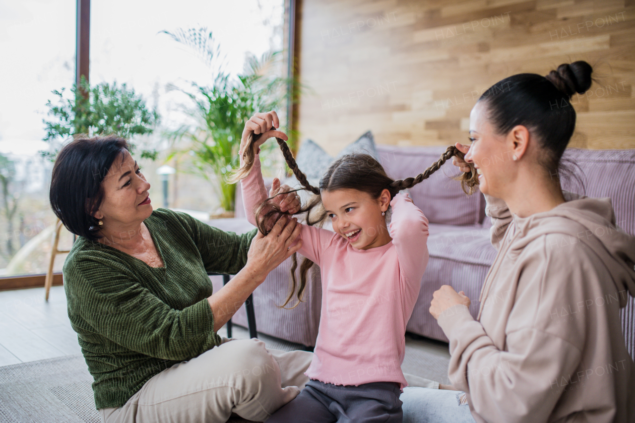 A happy little girl sitting on floor, her mother and grandmother making her plaits at home.