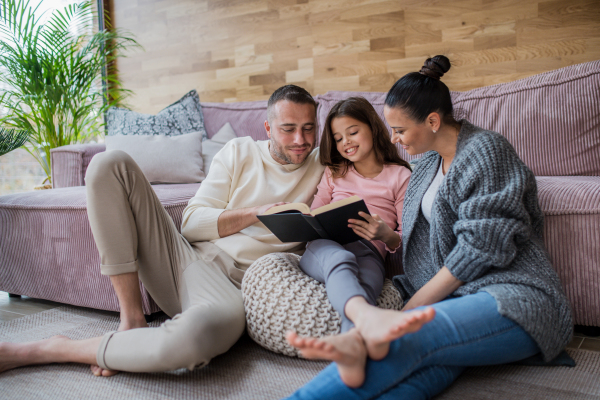 A happy little girl with mother and father sitting on floor and reading book at home