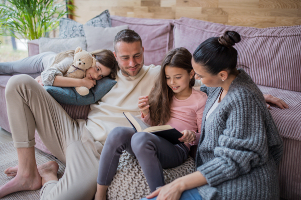 Two happy sisters with a mother and father sitting on floor hugging and reading book at home