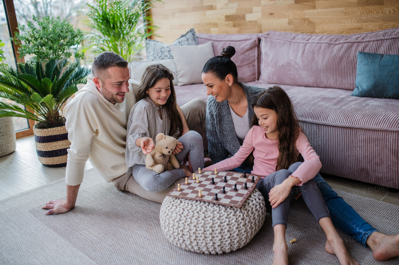 Two happy sisters with a mother and father sitting on floor and playing chess together at home