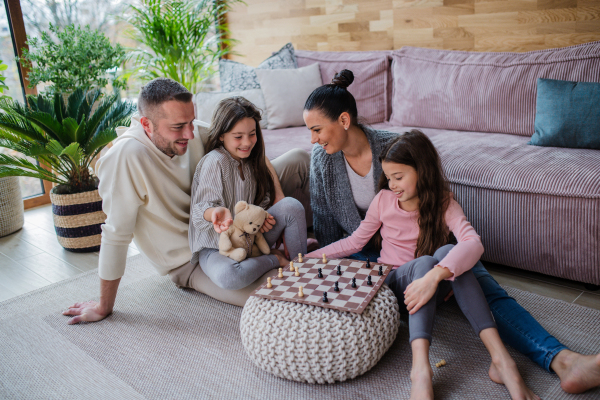 Two happy sisters with a mother and father sitting on floor and playing chess together at home