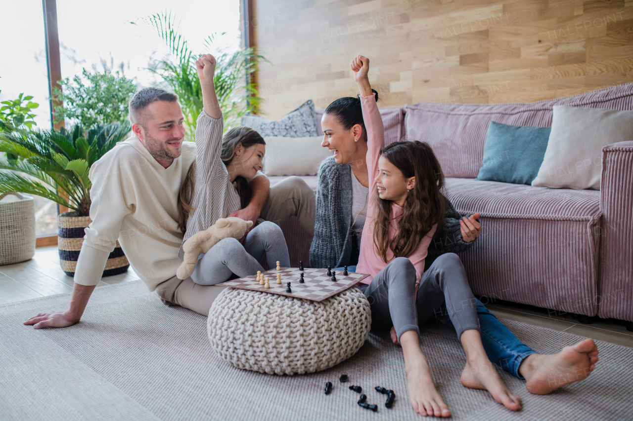 Two happy sisters with a mother and father sitting on floor and playing chess together at home