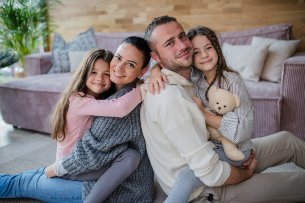 Two happy sisters with a mother and father sitting on floor hugging and looking at camera at home