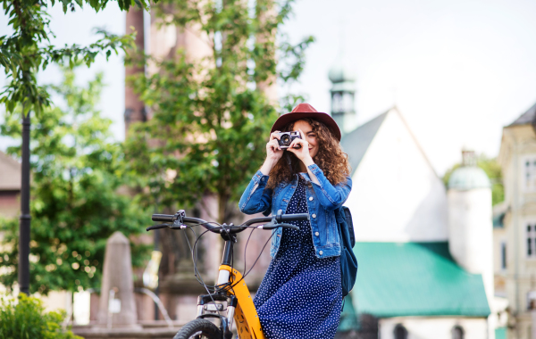 Beautiful young woman tourist traveller with electric scooter and camera in small town, taking photos.