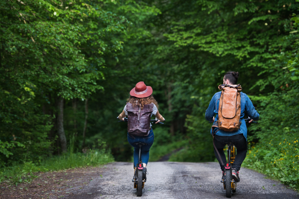 Rear view of young tourist couple travellers with backpacks and electric scooters in nature.