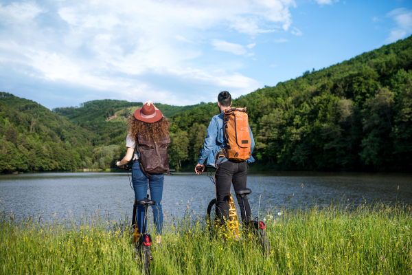 Rear view of young tourist couple travellers with electric scooters in nature, standing by lake.