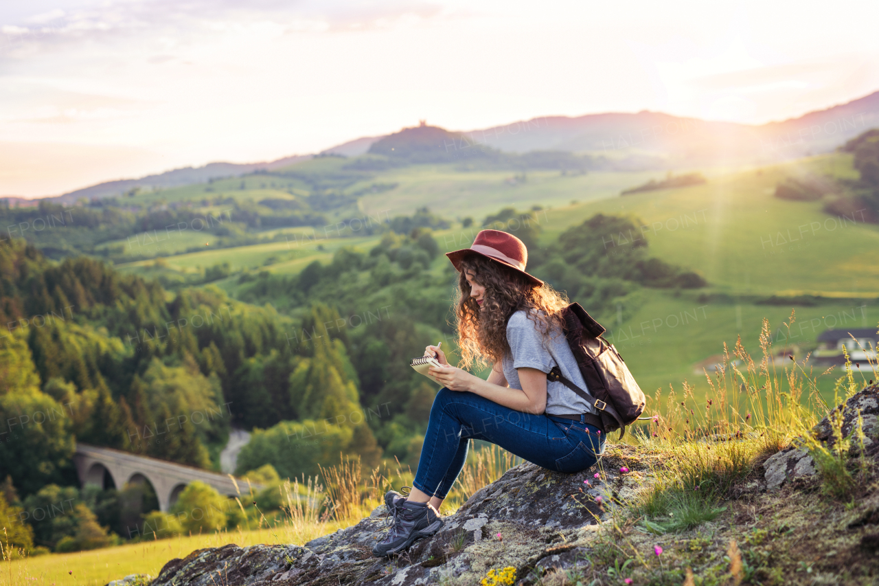 A young tourist woman traveller with backpack sitting in nature, writing notes.