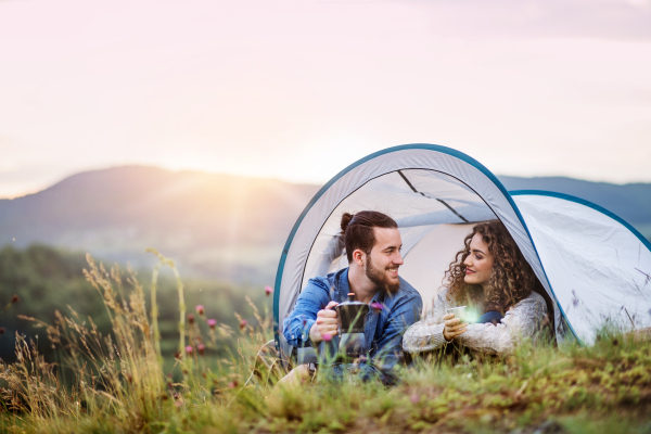 A young tourist couple travellers with tent shelter sitting in nature, drinking coffee.