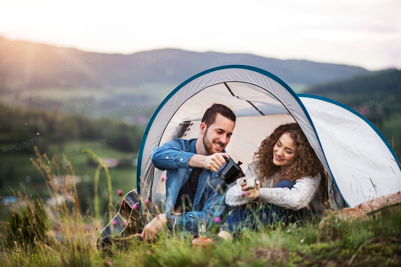 A young tourist couple travellers with tent shelter sitting in nature, drinking coffee.