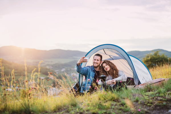 A young tourist couple travellers with tent shelter sitting in nature at sunset, taking selfie.