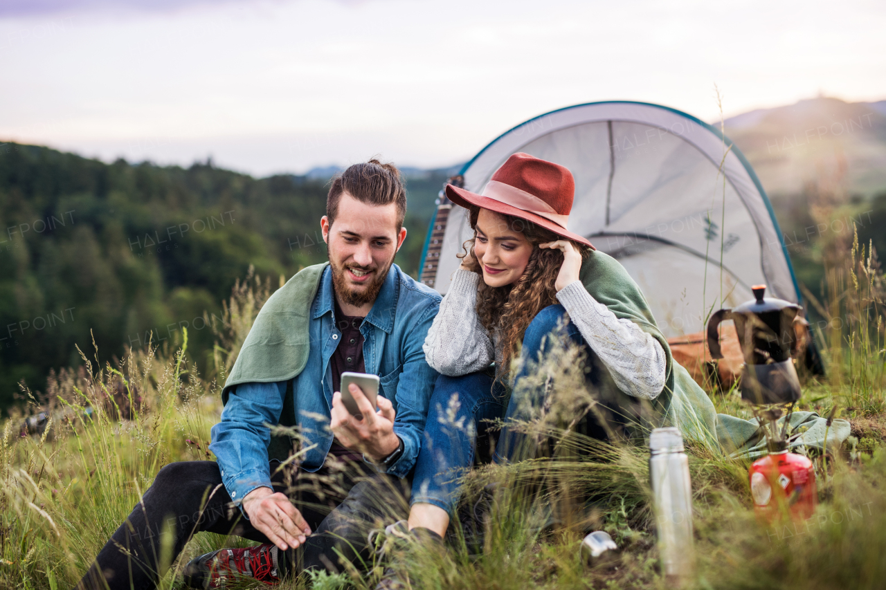 Young tourist couple travellers with tent shelter sitting in nature in the evening, using smartphone.