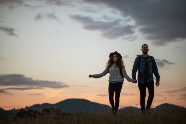 Young tourist couple travellers with backpacks hiking in nature at dusk, holding hands.