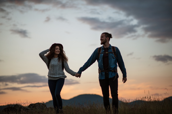 Young tourist couple travellers with backpacks hiking in nature at dusk, holding hands.