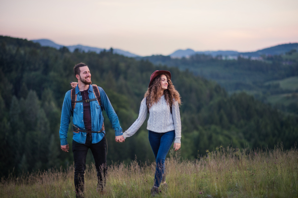 Young tourist couple travellers with backpacks hiking in nature at dusk, holding hands.