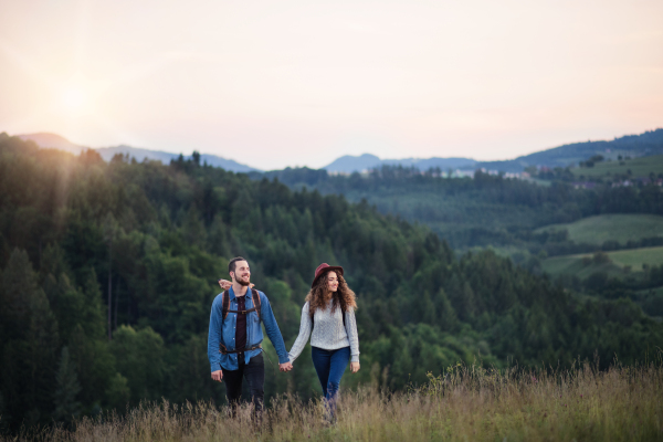 Young tourist couple travellers with backpacks hiking in nature at dusk, holding hands.